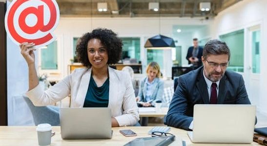 woman and man sitting at a desk with laptops, woman is holding up the @ sign and smiling