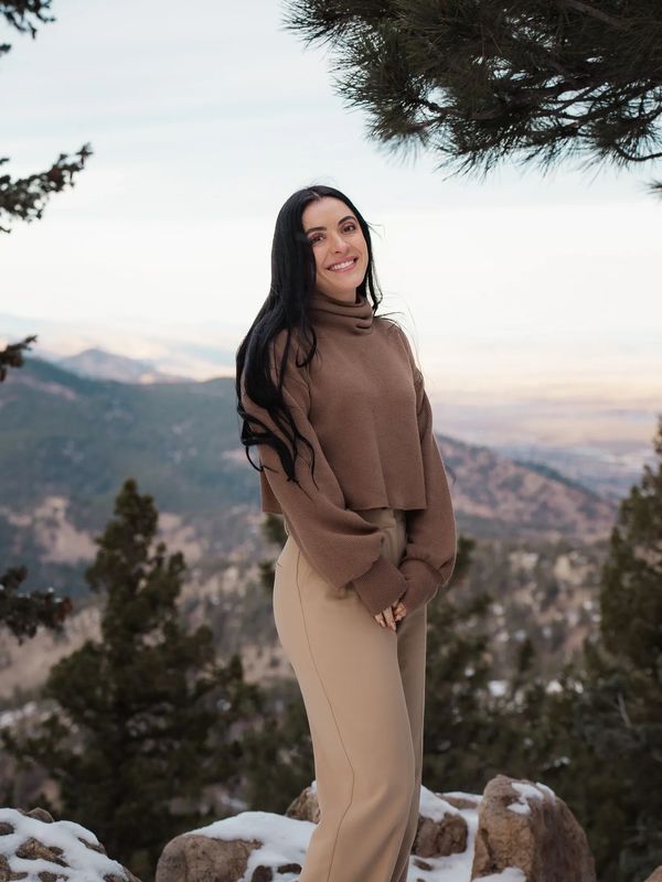 Smiling woman in brown sweater and beige pants stands on snowy mountain trail.