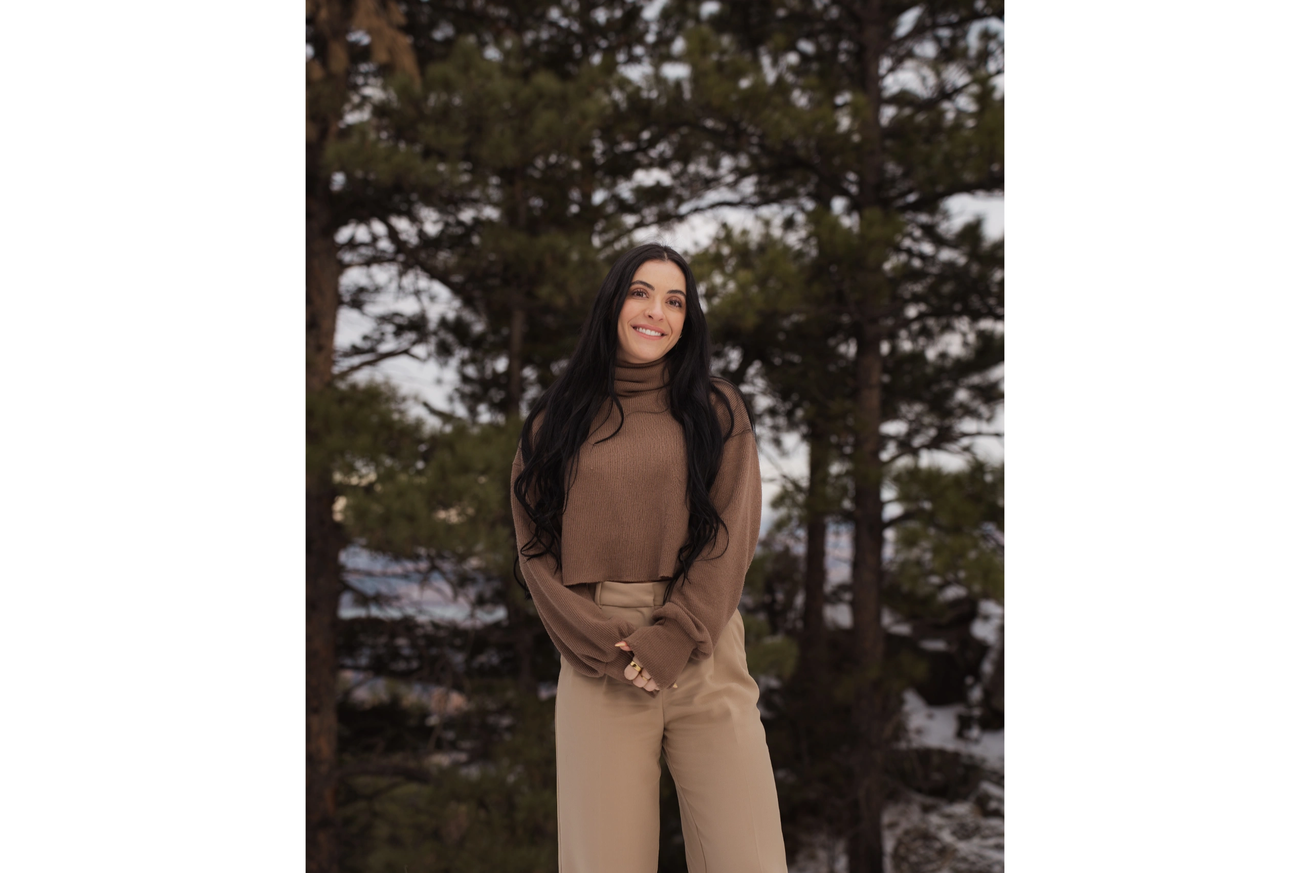 Smiling woman in a brown sweater and beige pants standing outdoors among trees.