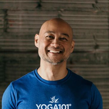 Smiling bald man in a blue YOGA101 shirt against a dark background.