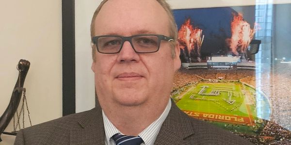 Man in suit and tie standing indoors with framed sports photo behind him.