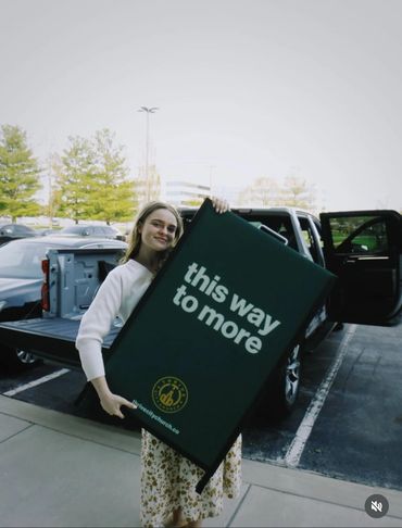 A smiling woman holding a sign that says 'this way to more' near a pickup truck.
