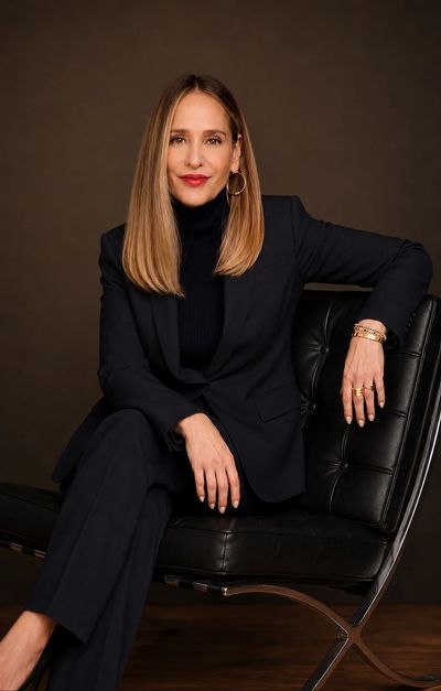 Confident woman in a dark suit sitting on a modern chair against a brown background.