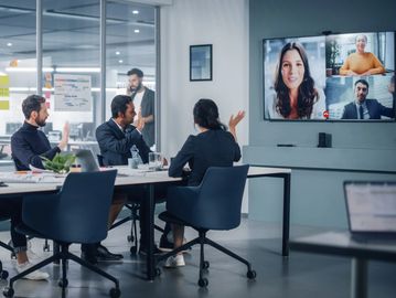 Colleagues in a hybrid meeting waving to remote participants on a video call.
