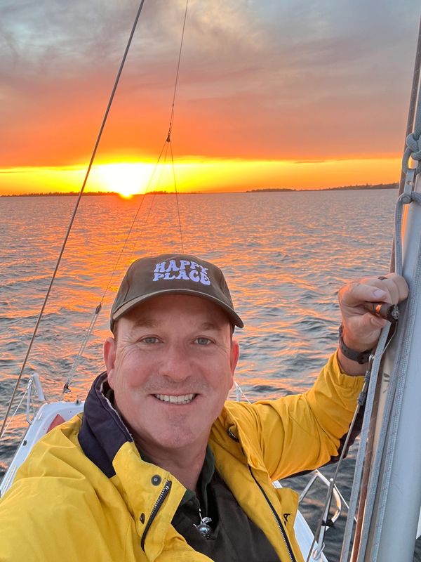 Man in yellow jacket and cap smiling on a boat at sunset.