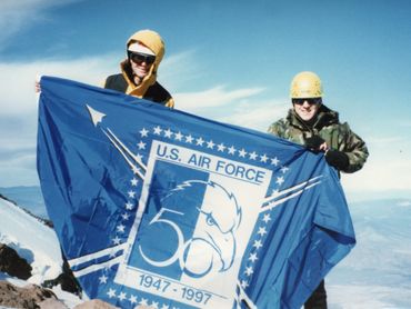Two climbers hold a U.S. Air Force 50th anniversary flag on a snowy peak.