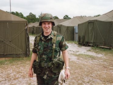 Soldier in camouflage uniform smiling near military tents.
