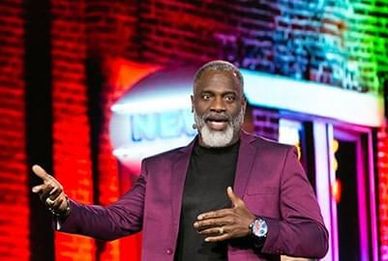 Man with gray beard speaking on stage wearing a purple blazer.