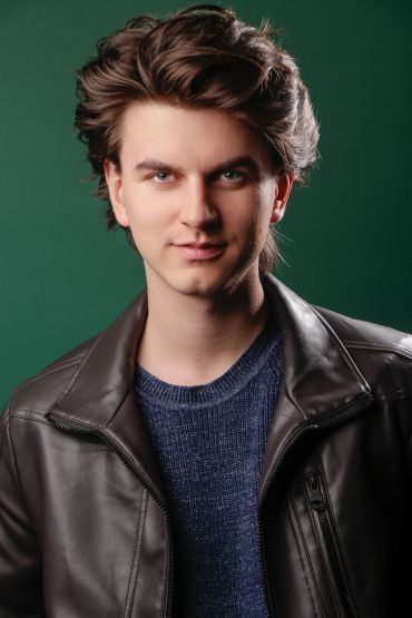 Young man with styled hair wearing a leather jacket and blue sweater.