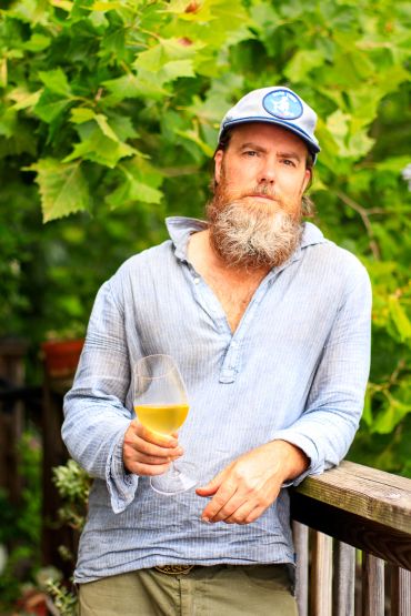 Man with beard holding a glass of white wine outdoors.