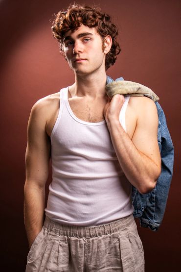 Young man with curly hair wearing a white tank top and holding a jacket.
