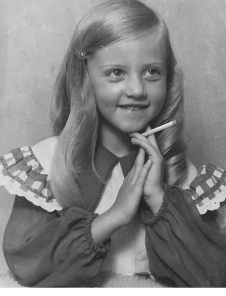 Young girl with long blond hair holding a cigarette and smiling.