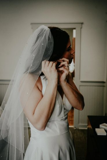 Bride adjusting earrings, wearing a white wedding dress and veil indoors.