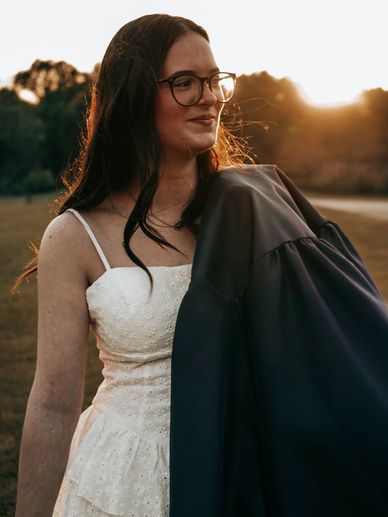 Woman in white dress holding a dark jacket, smiling at sunset.