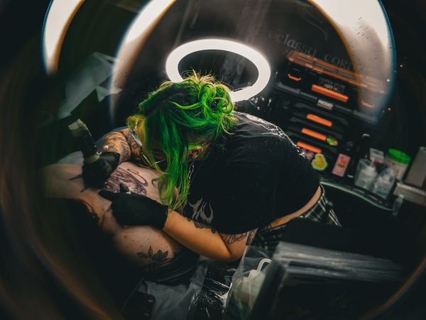 Tattoo artist with green hair working on a client's back tattoo under a ring light.