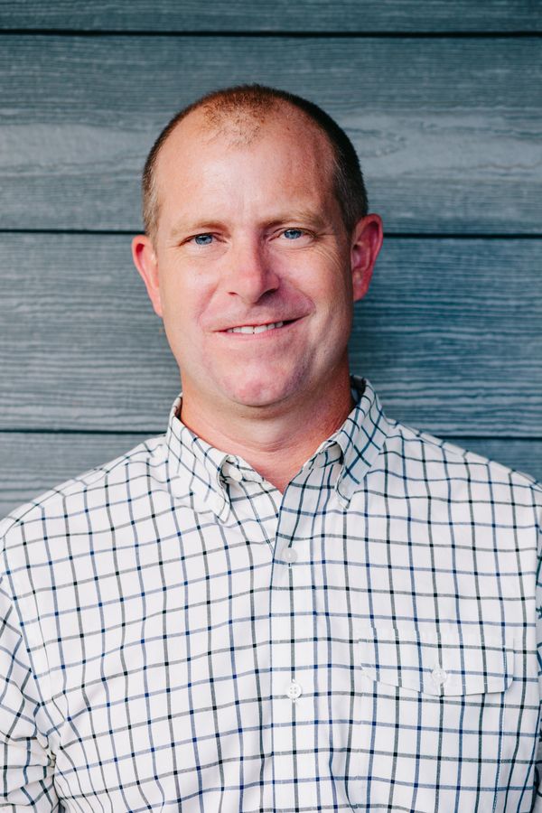 Smiling man in a checkered shirt stands against a wooden wall.