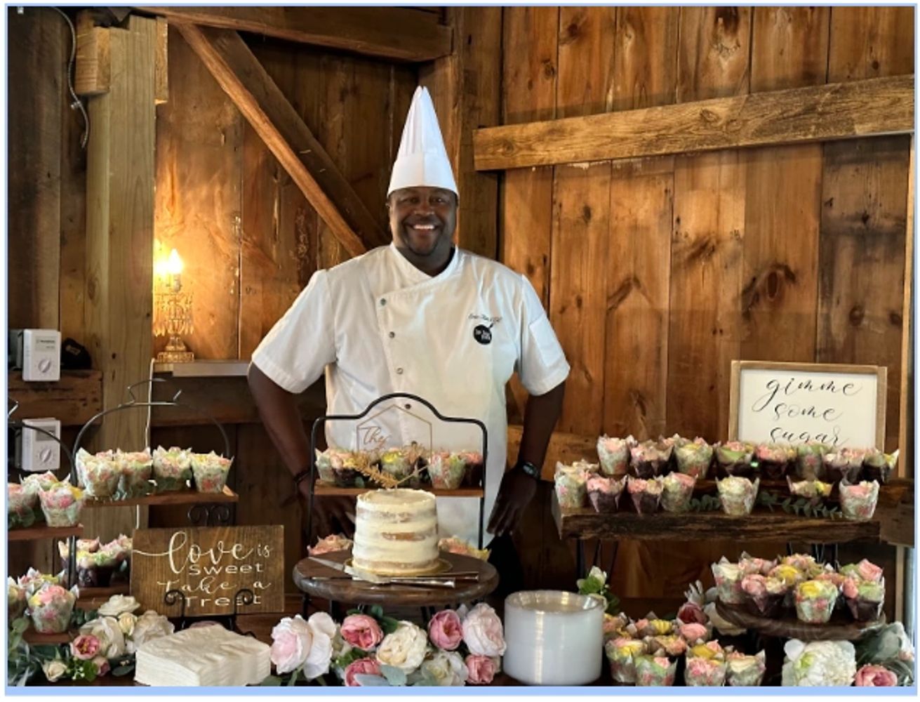 Smiling chef stands behind a dessert table with cupcakes and a cake in a rustic setting.