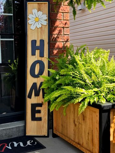 Wooden HOME sign with a daisy next to green ferns on a porch.