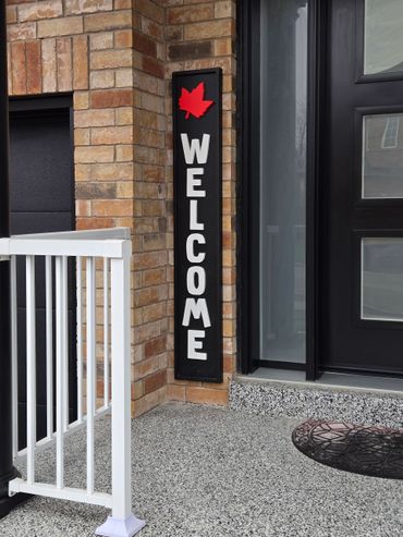Vertical welcome sign with a red maple leaf by a modern black door.