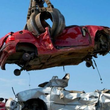 A crane lifting a crushed red car over a pile of crushed vehicles at a scrapyard.
