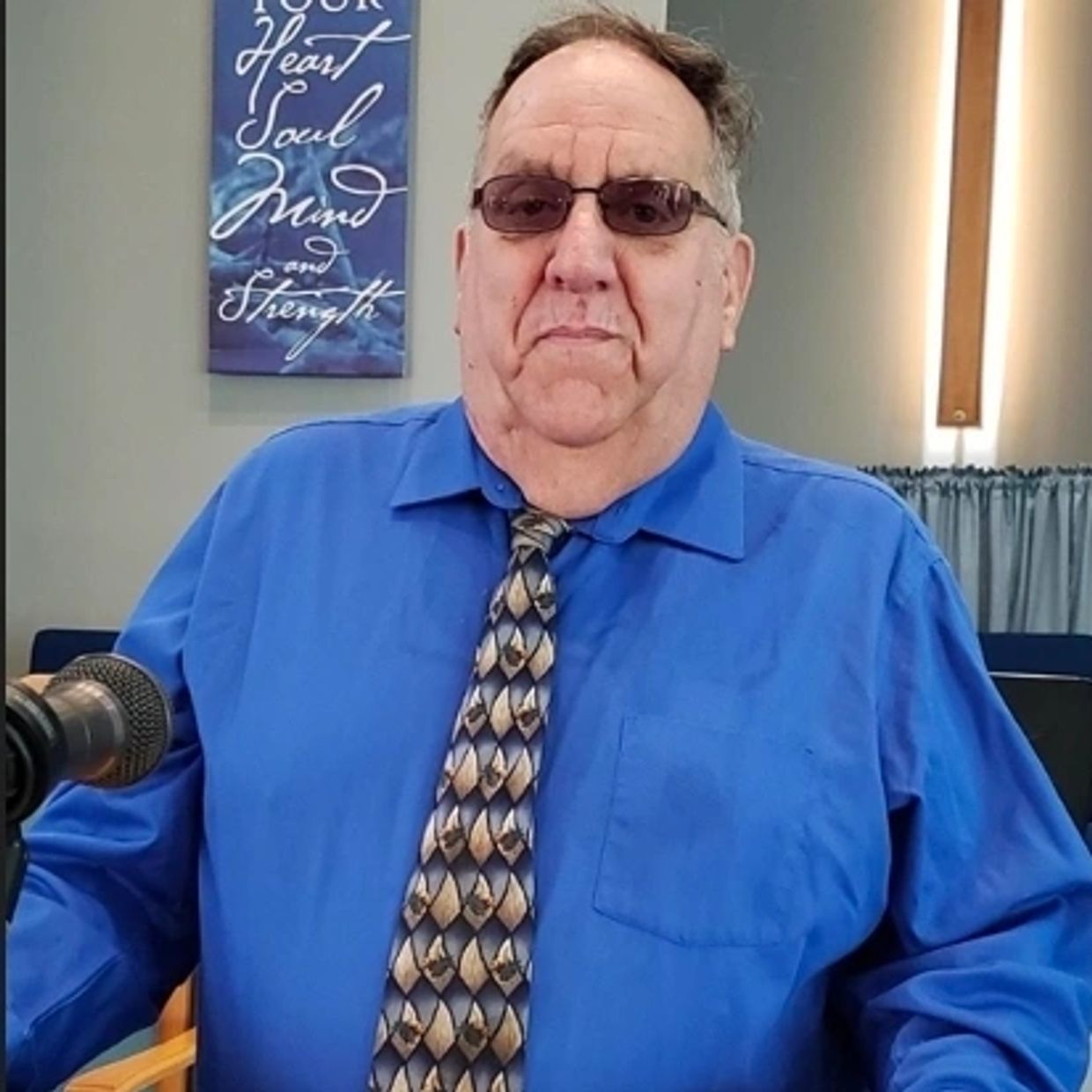 Man in blue shirt and patterned tie sitting in front of a microphone.