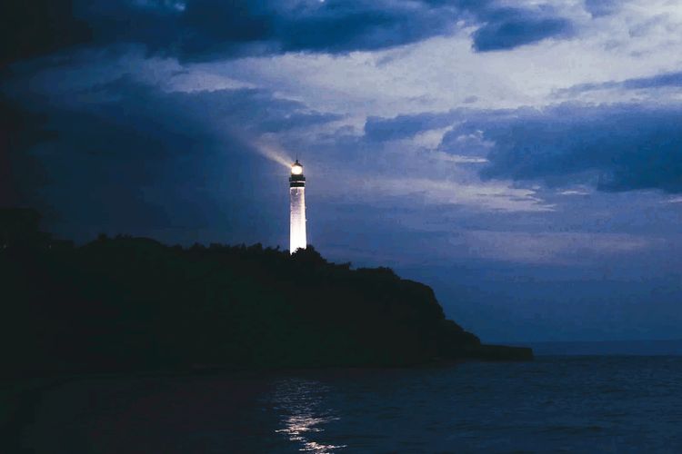 A lighthouse shines its beam over a dark sea under a cloudy night sky.