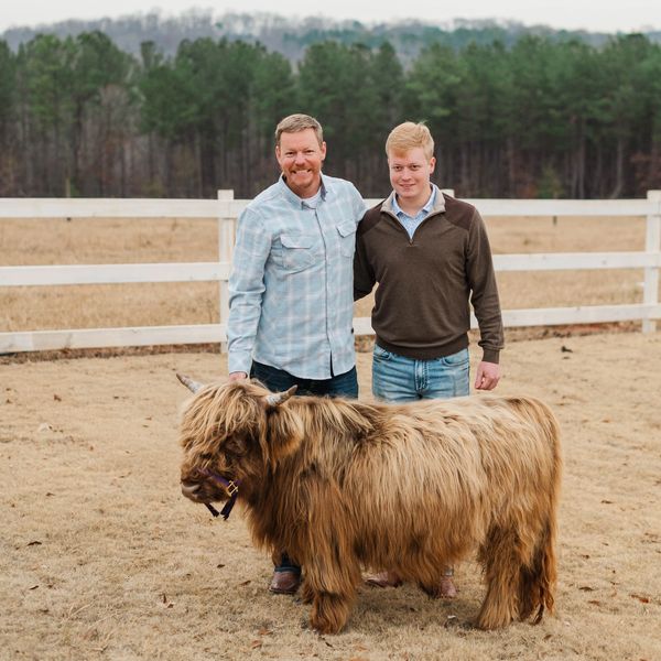 Two men standing outdoors with a shaggy Highland cow in a fenced field.