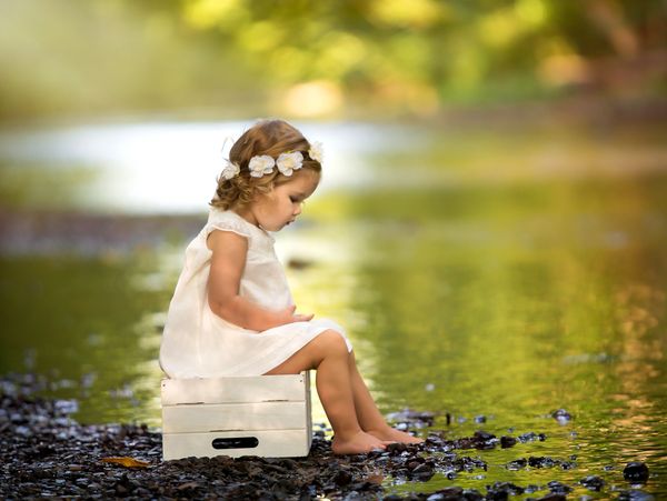 A young girl in a white dress sitting on a crate by a calm river.