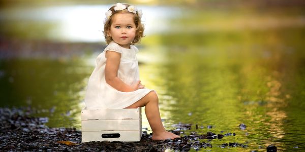 A young girl in a white dress sitting on a white crate near a water body.