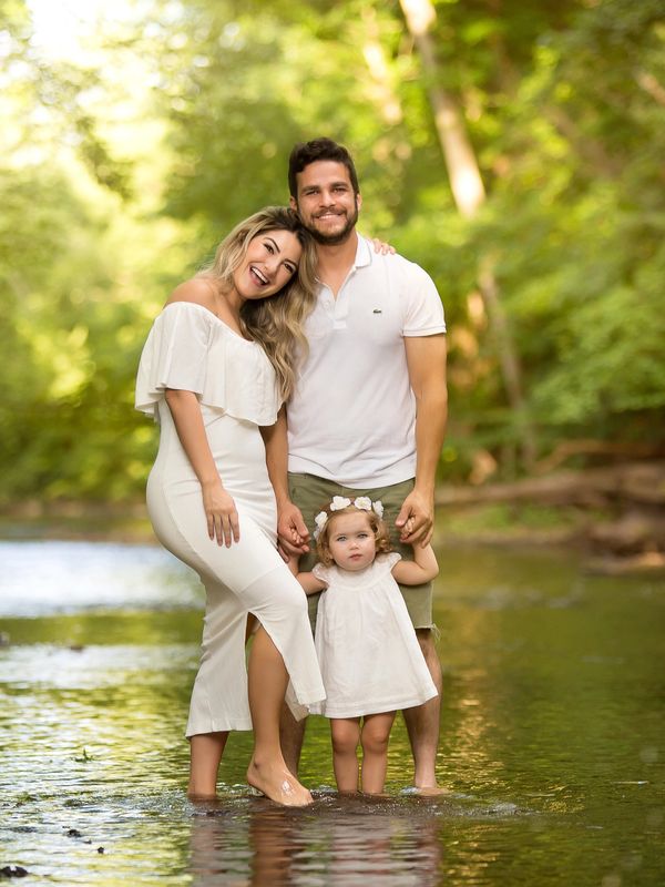 A happy family of three standing barefoot in a shallow river, surrounded by lush greenery.