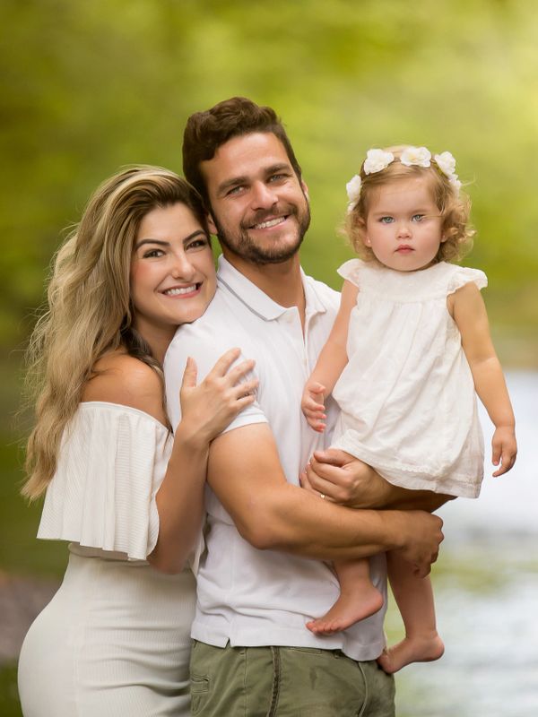 Smiling family of three in white outfits outdoors by water.