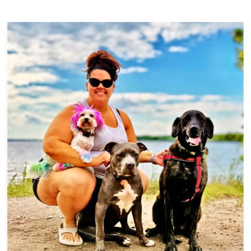 Woman with three dogs by a lake, two large and one small in colorful attire.