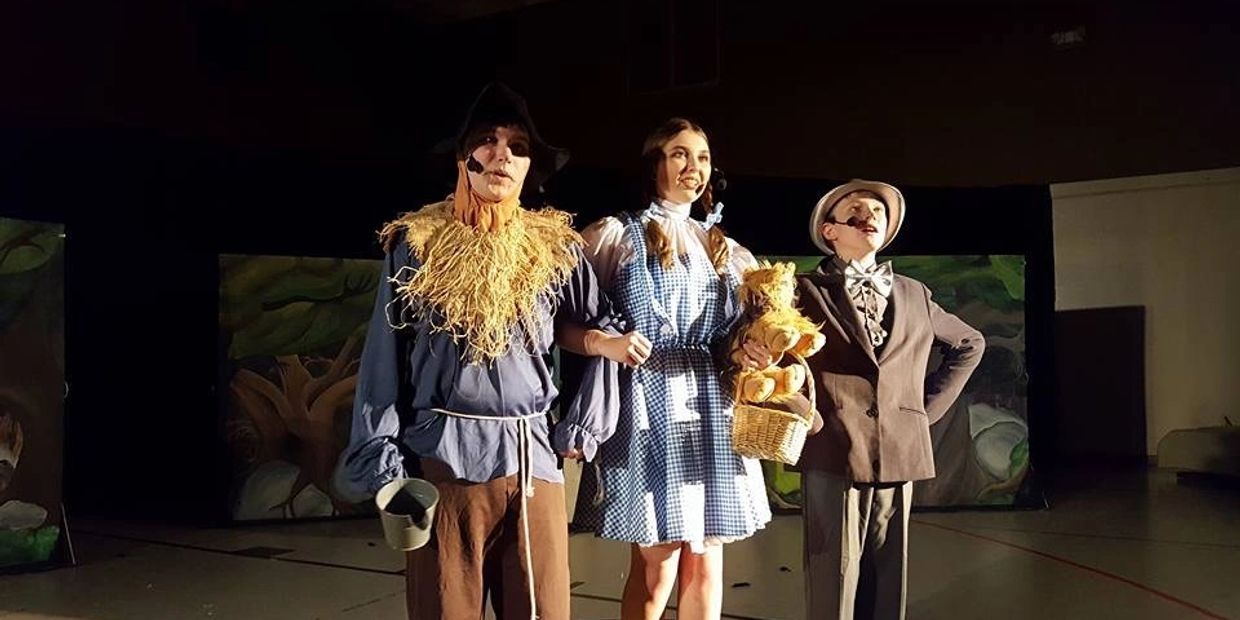 Three actors in Wizard of Oz costumes on stage, spotlighted.