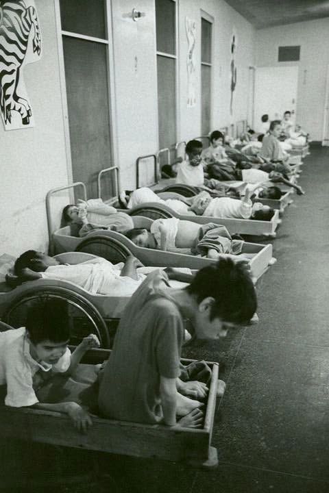 Children with disabilities resting in small wooden beds in a hospital corridor.