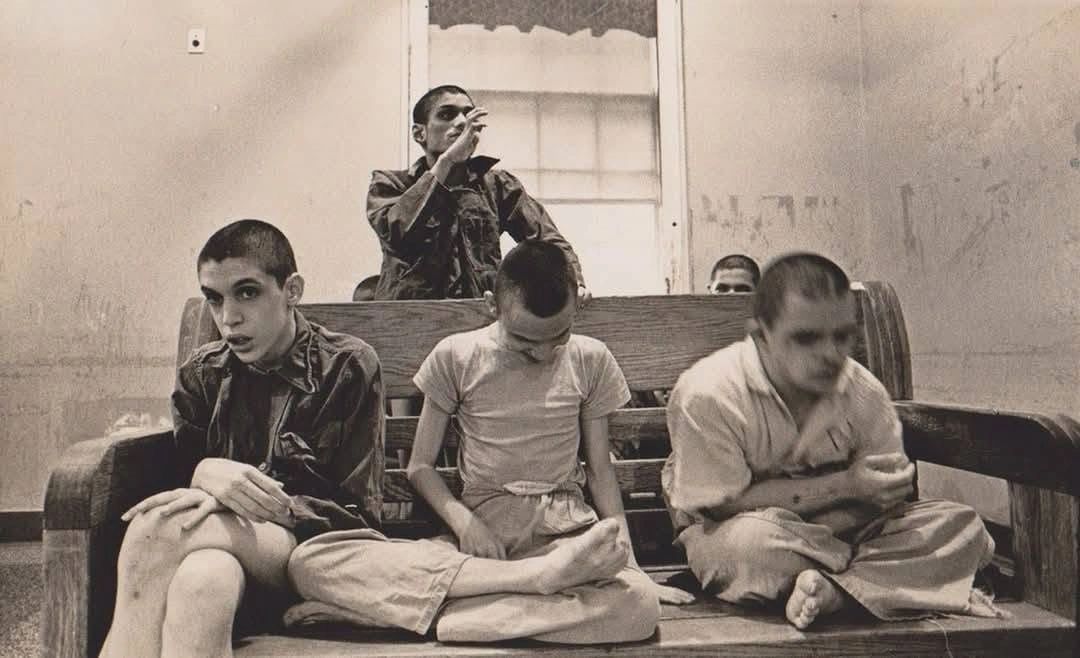 Several young men with shaved heads sitting and standing in a sparse room with a wooden bench.