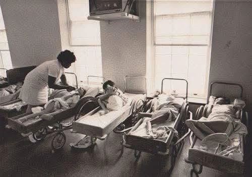 Nurse tending to children in a hospital ward with vintage bassinets.