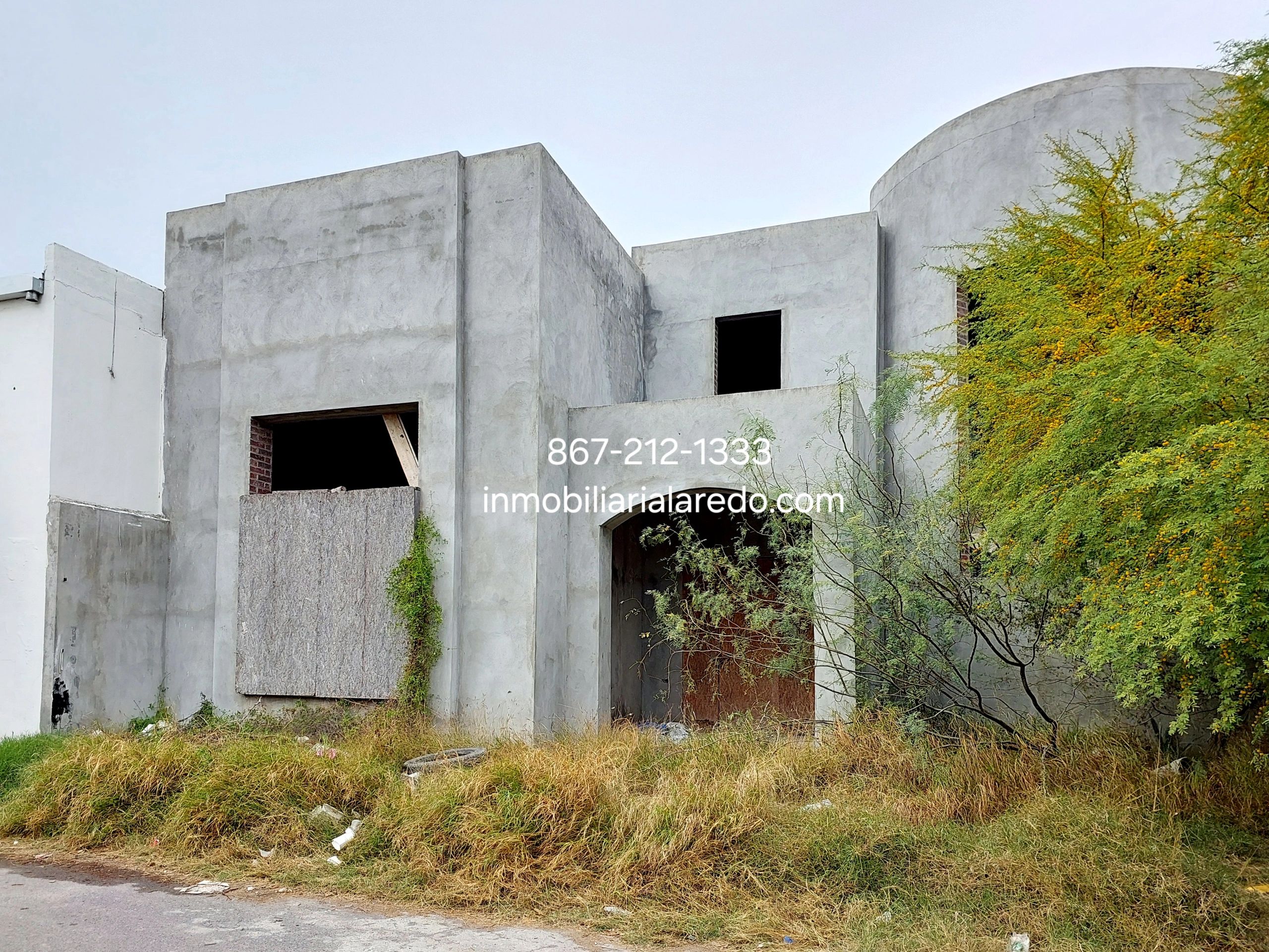 Unfinished concrete house with boarded windows and overgrown grass.