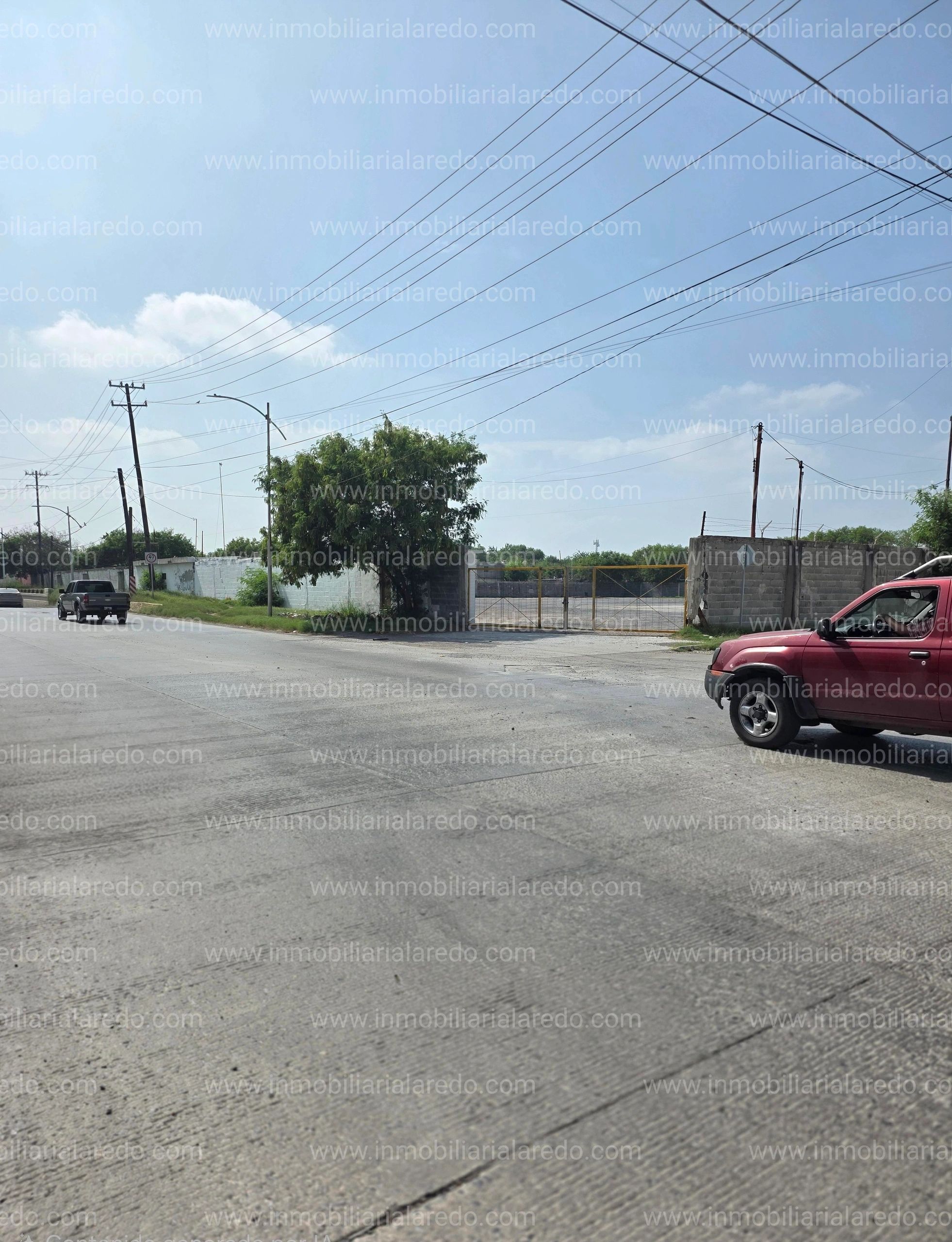 Street scene with vehicles, trees, power lines, and a gated entrance under a clear sky.
