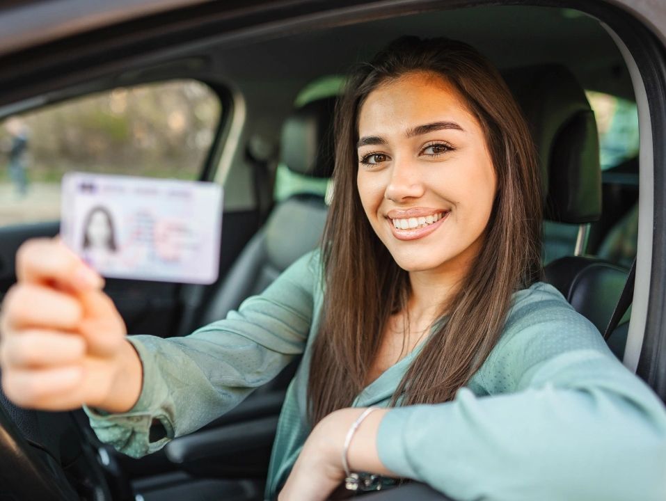 Woman in car holding up a driver's license and smiling.