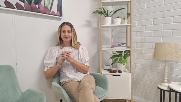A woman sitting on a mint green chair in a cozy, plant-filled room.