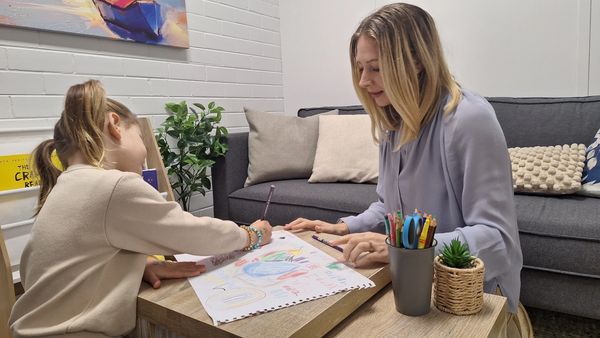 A woman and girl drawing together at a wooden table in a cozy living room.