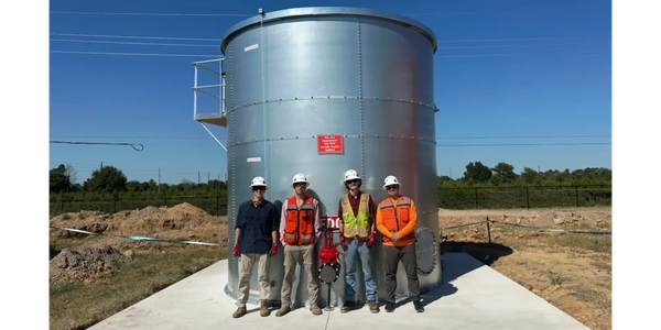 Four construction workers stand in front of a large metal storage tank on a concrete platform.