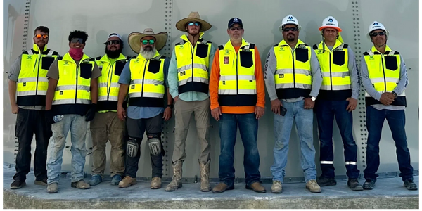 Group of construction workers in high-visibility vests posing against a metal wall.