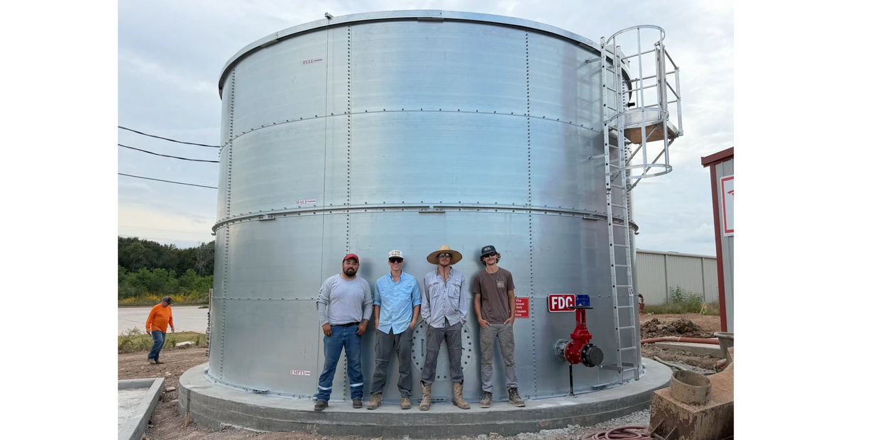 Four men standing in front of a large metallic water tank outdoors.