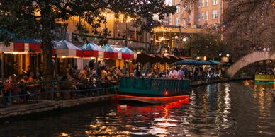 Boat cruising on a lively riverwalk with outdoor diners at sunset.
