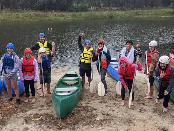 Group of people in life vests with paddles and a green canoe on a riverbank.