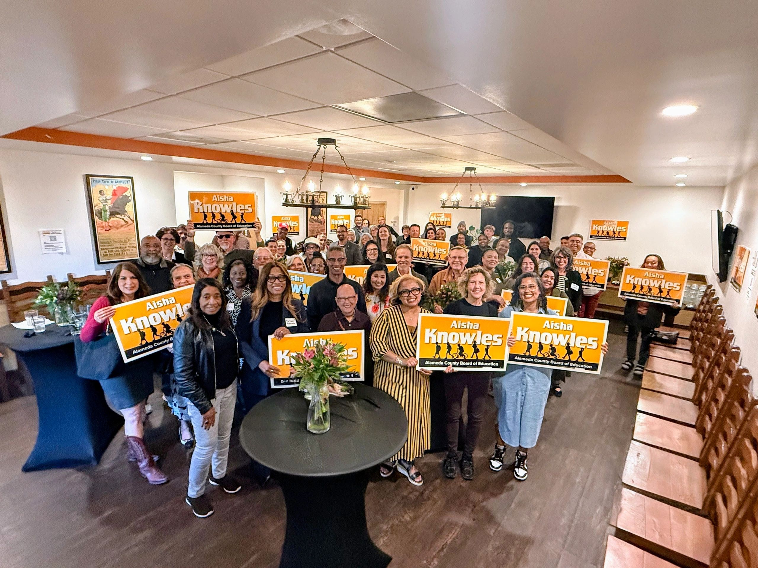 Large group holding "Aisha Knowles" campaign signs indoors at an event.