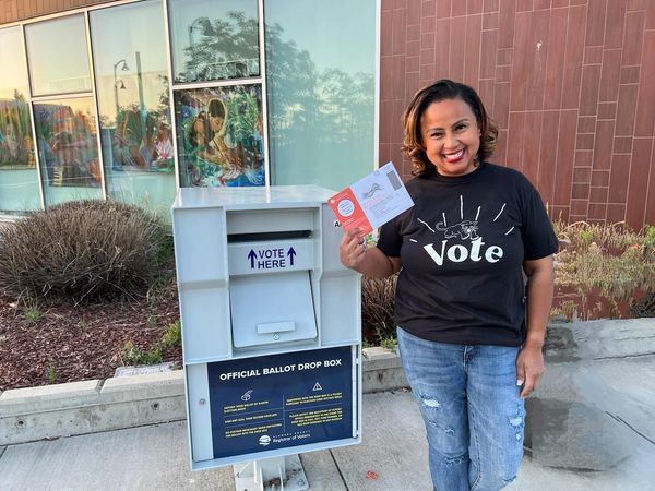 Woman smiling while holding a ballot near an official ballot drop box.