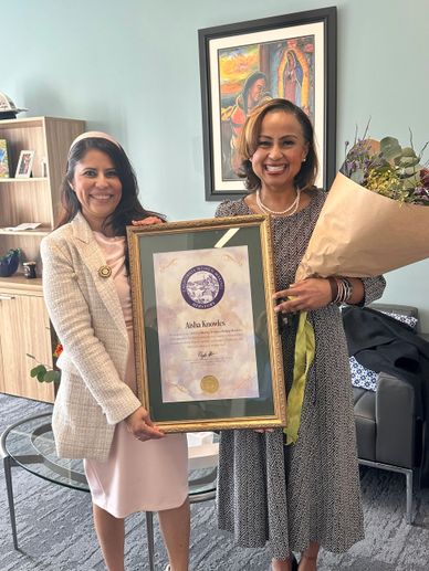 Two women smiling, one holding a framed certificate and the other holding a bouquet of flowers.