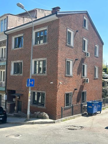 Three-story brick building on a sunny street corner with air conditioners and blue trash bins.